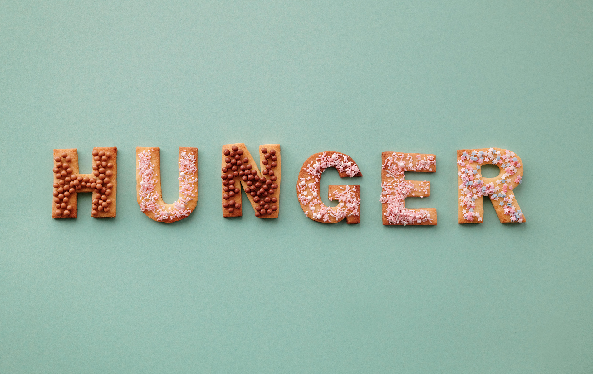 Das Wort „HUNGER“ besteht aus bunten, mit Zuckerguss verzierten Keksen auf einem hellgrünen Hintergrund und steht symbolisch für Food Noise und ständige Gedanken an Essen.