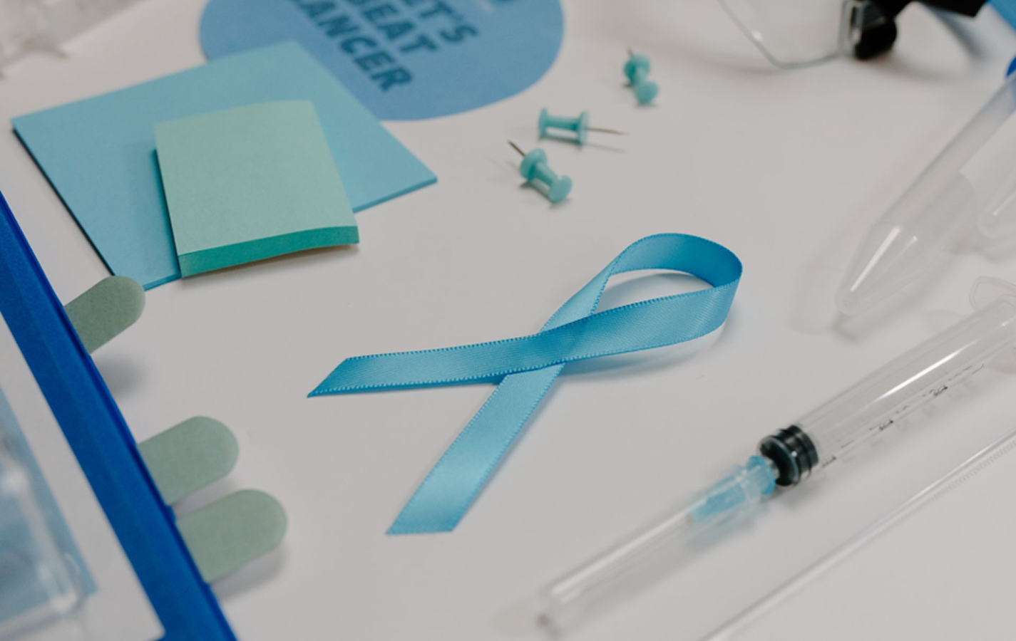 Das Krebs-Ribbon auf einem weißen Tisch mit Notizzetteln und Spritzen als Symbolbild für den Zusammenhang zwischen Übergewicht und Krebs. 